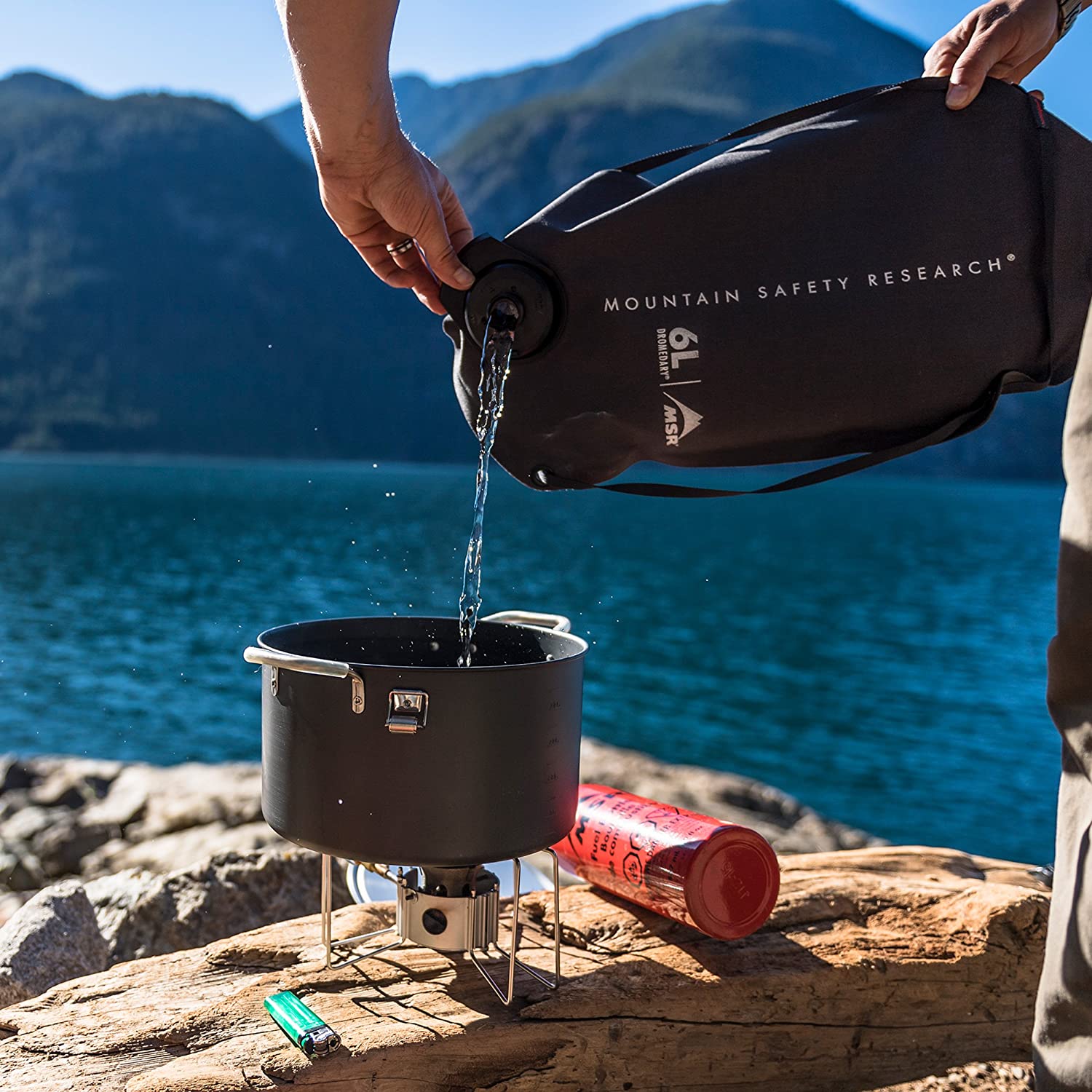 A person pouring filtered water into a camp stove pot from a Mountain Safety Research (MSR) rugged water storage bag. A large lake and hillside are shown in the distance.
