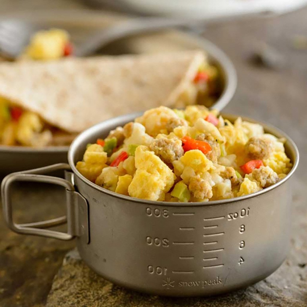 A pot of Mountain House Breakfast Skillet prepared freeze dried food, with measurements 100, 200, and 300 milimeters written on the side. In the background is a plate with a tortilla wrapped witht he Breakfast skillet makings.