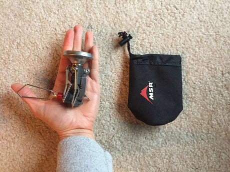 A person holding a MSR PocketRocket camp stove in the palm of their hand beside it's small black carrying case on a beige carpet.