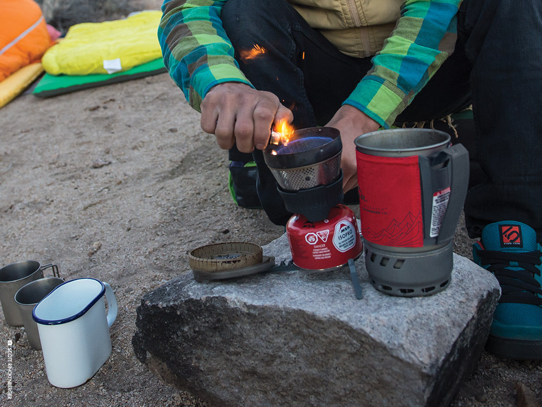 Someone sparking a camp stove with a lighter. A sleeping bag is in the background in yellow, 3 camping mugs sit by the stove.