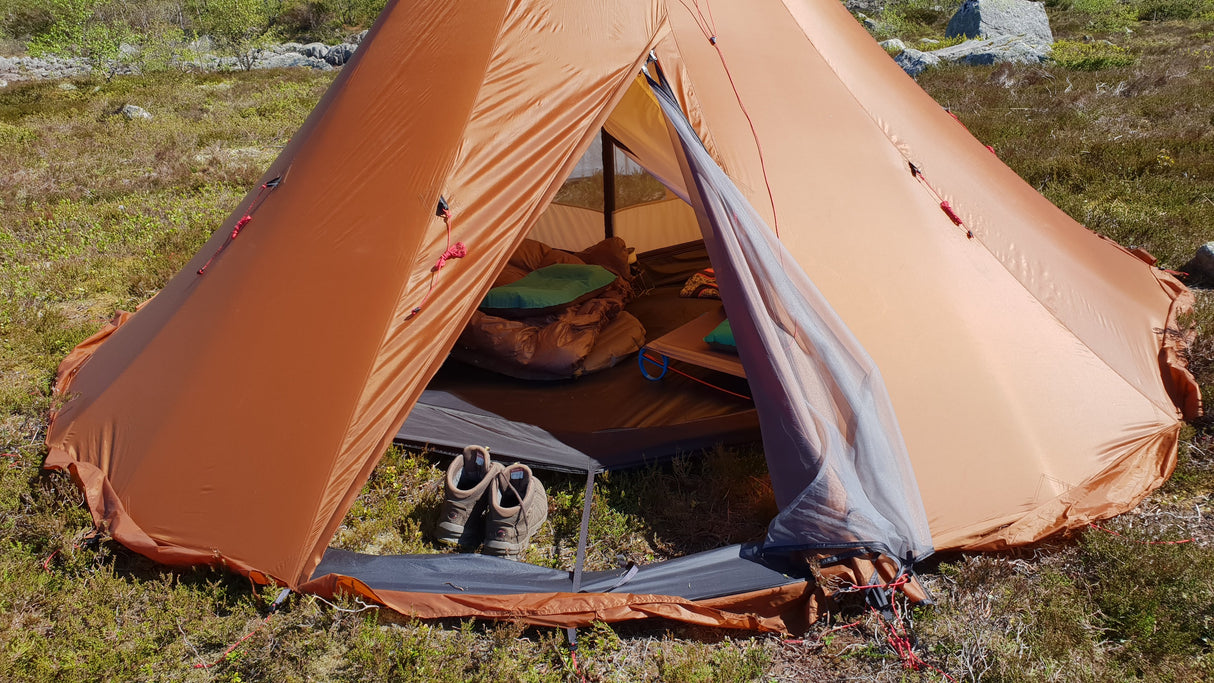 The NorTent Iavvo 4 Winter Hot tent with a persons hiking shoes, sleeping bag, and mattress in the middle of the inside of the tent. The tent is a copper brown colour with a grey bug mesh in the entry way door.