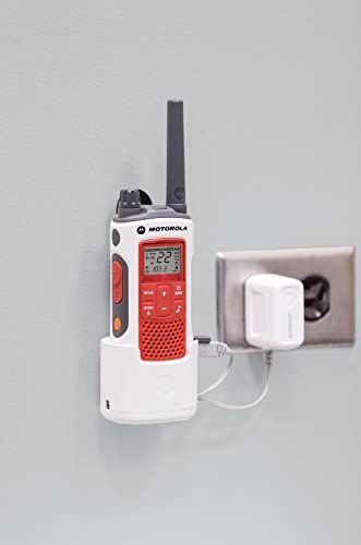 A Motorola Talkabout T480 holstered in it's charging dock and being charged next to an outlet on a wall. The walkie talkie is a white, red and black color. The charging dock is white and so is the wall charger.