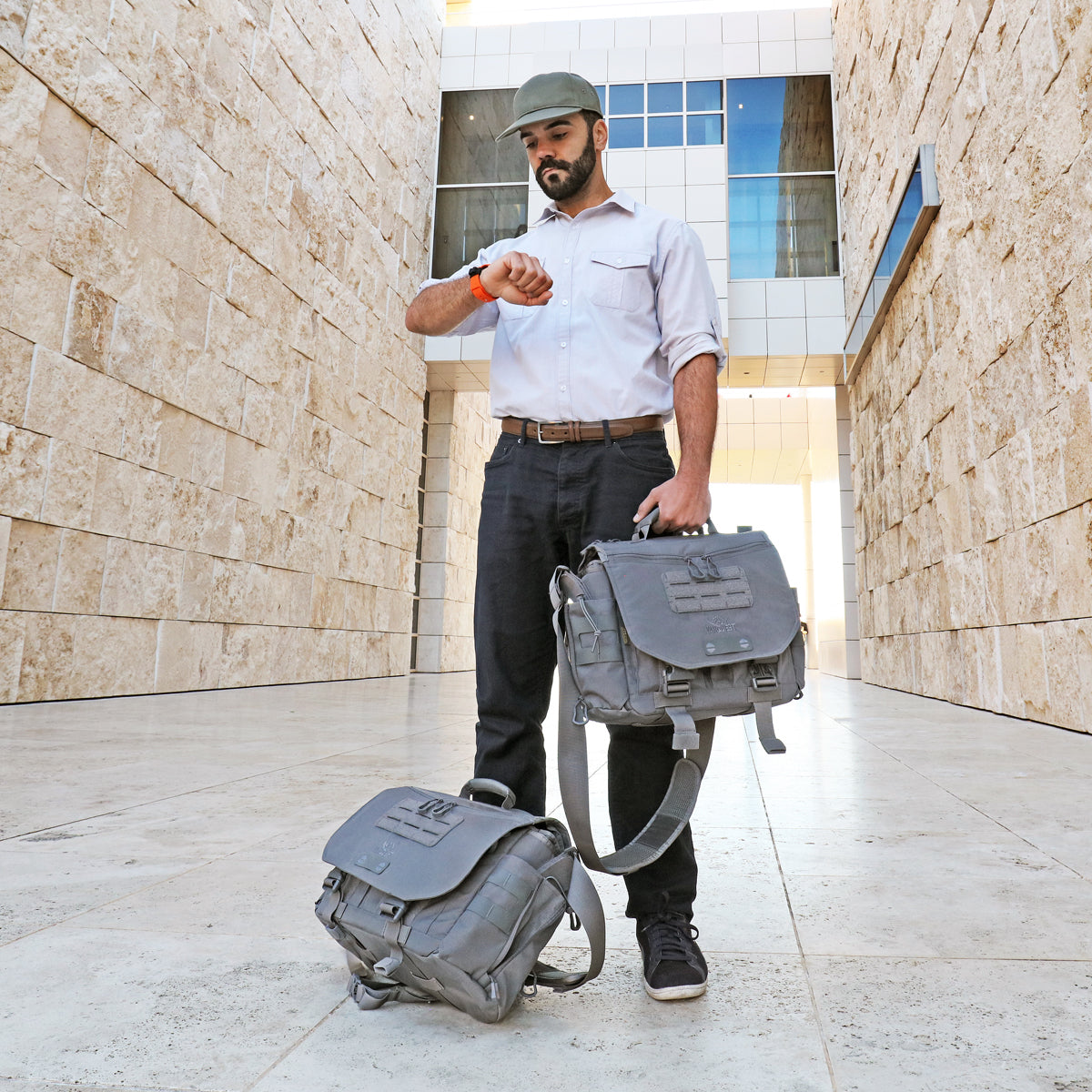 Traveler with two Gray Vanquest ENVOY-17 (Gen-4) Messenger Bags checking the time and awaiting a pickup to the airport outside a large marble stone building.