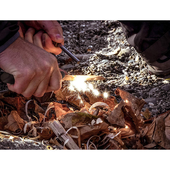 A man using a Morakniv bushcraft knife with a fire striker to start a fire on a pile of dry leaves and wood shavings on assorted rocks and debris.