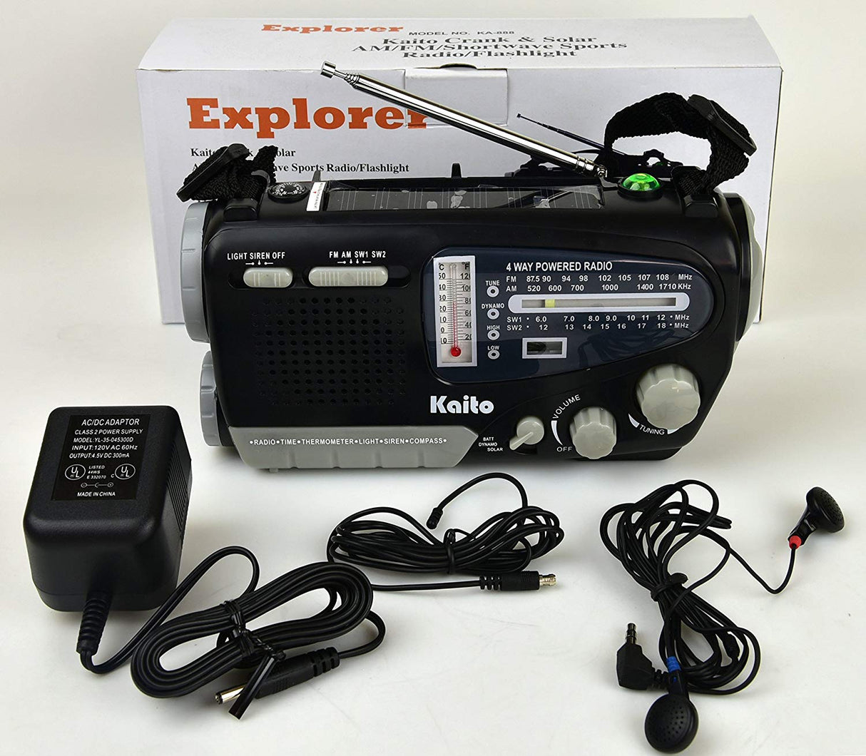 Explorer Kaito Ka888 emergency radio with am/fm tuner, volume knob, tunning knob, light siren knob, and SW1/SW2 frequencies. Beside are a pair of included black headphones, attenna for fm, and a charging block.