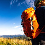 A UCO titanium spork hooked to an orange marmot backpack. A hiker wears the pack with spork walking through a field of tall grass. Rolling green hills and blue sky populate the background.