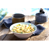 A white and blue coloured bowl of Happy Yak prepared chicken blanquette and egg noodles is set on a picnic table with a camping mug and hot kettle beside it.