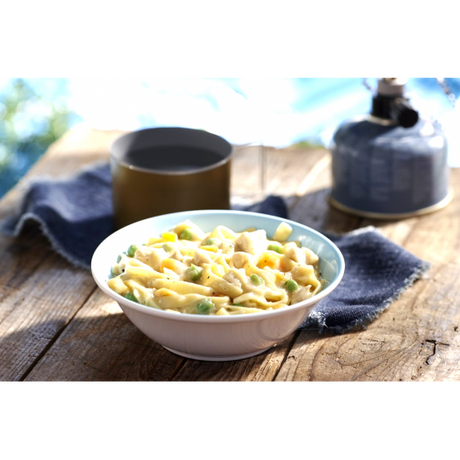 A white and blue coloured bowl of Happy Yak prepared chicken blanquette and egg noodles is set on a picnic table with a camping mug and hot kettle beside it.