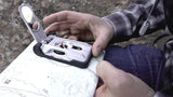 A man in a plaid shirt with his backpack straps hanging, holds the Brunton TruArc 20 compass on a map, to navigate his surroundings and better understand his real world location.