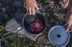 A person stowing away the contents of a Mountain Safety Research Windburner Group stove system in it's cooking pot. The red propane tank and cooking stove both fit in the pot. A rock and grass covered ground are below.