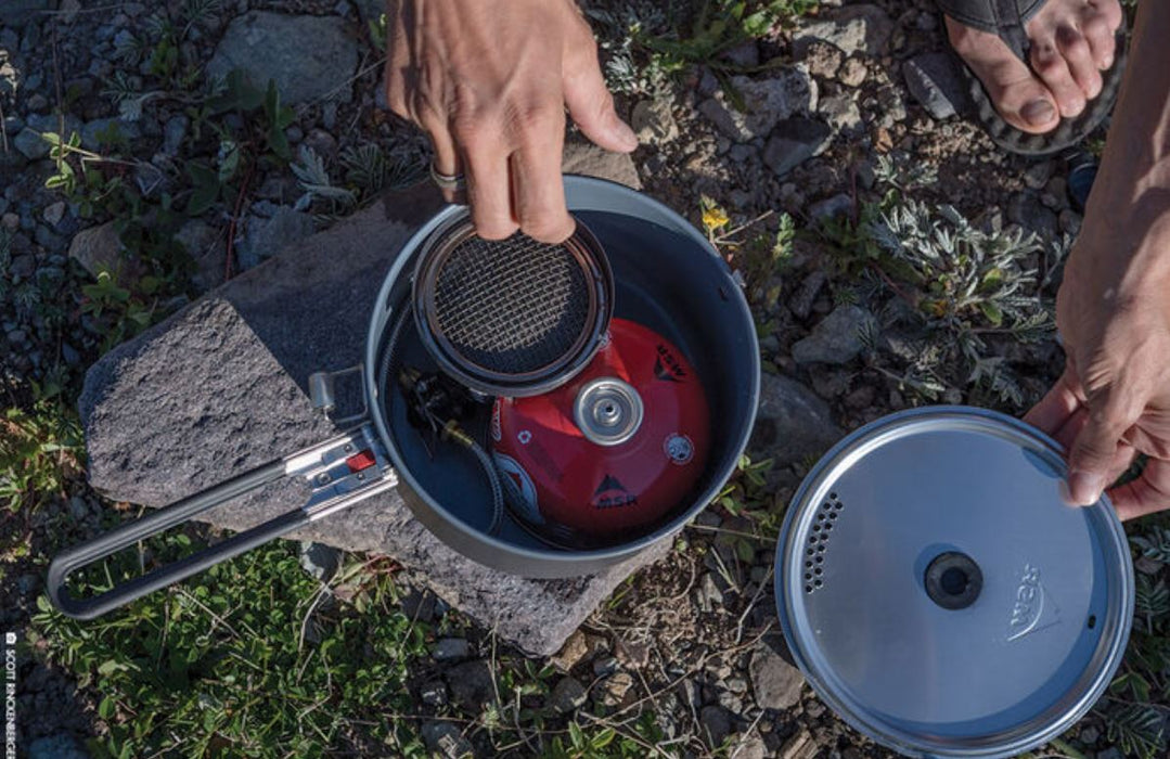 A person stowing away the contents of a Mountain Safety Research Windburner Group stove system in it's cooking pot. The red propane tank and cooking stove both fit in the pot. A rock and grass covered ground are below.
