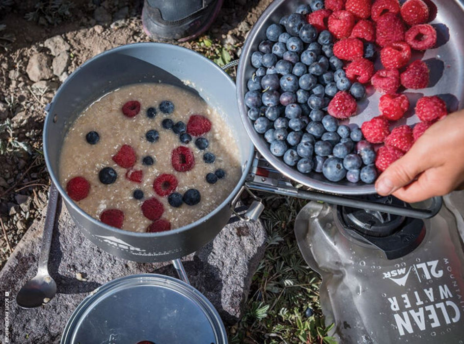 A person holding a titanium bowl of fresh blueberries and raspberries putting them into a titanium pot of oatmeal. A 2L rugged water bag from MSR is shown beside the bowl and everything is on a rocky covered ground.