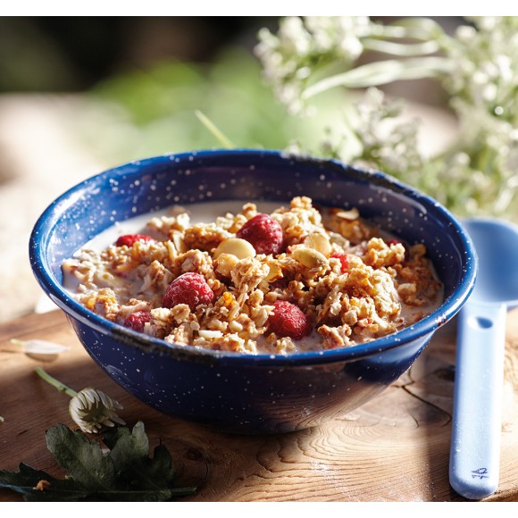 A white spotted navy blue bowl of Happy Yak Granola Raspberry and Vanilla with milk. A plastic camping spoon is beside the bowl, both are on a tree stump with white flowers beside.