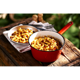 A red camping cooking pot full of Happy Yak's Mom's Tomatoe and Cheese Macaroni, with a white bowl of the food laid on a wood crate in the outdoors.