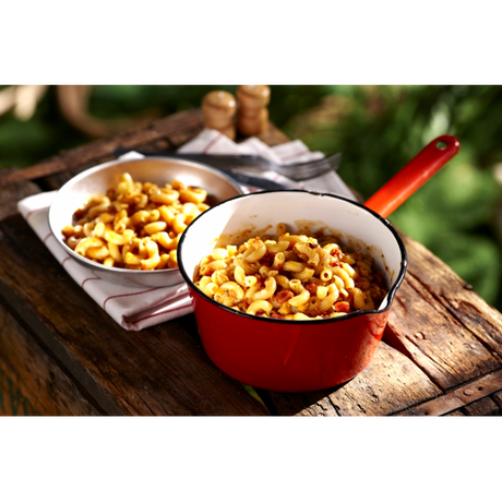 A red camping cooking pot full of Happy Yak's Mom's Tomatoe and Cheese Macaroni, with a white bowl of the food laid on a wood crate in the outdoors.