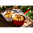 A red camping cooking pot full of Happy Yak's Mom's Tomatoe and Cheese Macaroni, with a white bowl of the food laid on a wood crate in the outdoors.