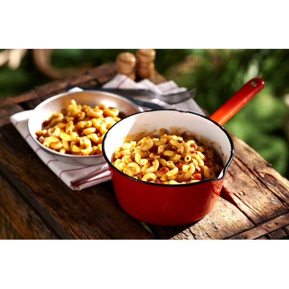 A red camping cooking pot full of Happy Yak's Mom's Tomatoe and Cheese Macaroni, with a white bowl of the food laid on a wood crate in the outdoors.