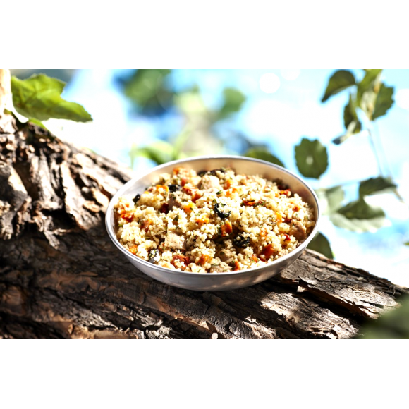 A silver stainless steel camping bowl full of the Happy Yak mediterranean Pork dish on a large tree branch wth green leaves surrounding it.