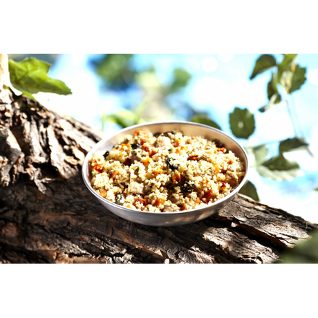 A silver stainless steel camping bowl full of the Happy Yak mediterranean Pork dish on a large tree branch wth green leaves surrounding it.