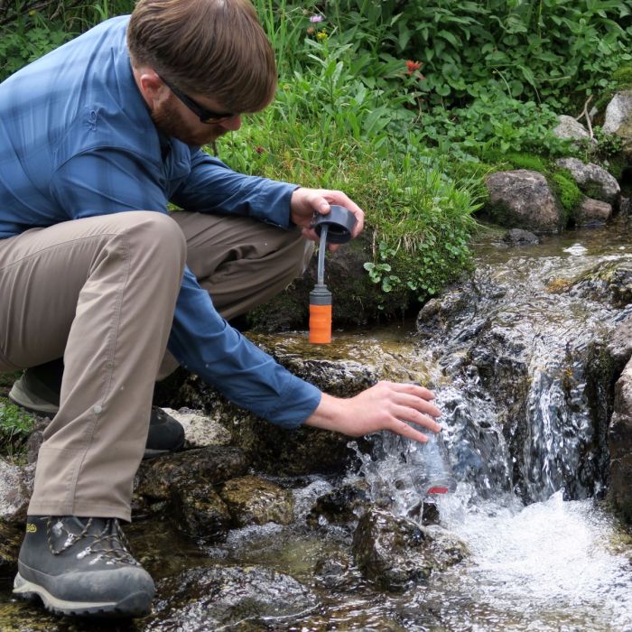 A man wearing a blue plaid shirt, black sunglasses and kahki hiking pants filling a Muv Nomad water bottle up in a river. He holds the water filter and bottle cap in his other hand while filling it.