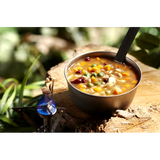 A camping bowl of Happy Yak Chicken Orzo Freeze Dried Soup is shown heated up by a camp stove torch on a tree stump outdoors.