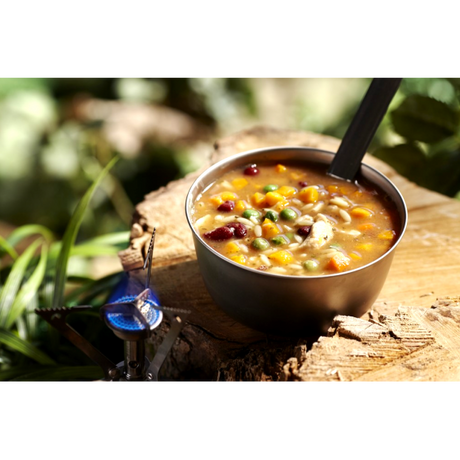 A camping bowl of Happy Yak Chicken Orzo Freeze Dried Soup is shown heated up by a camp stove torch on a tree stump outdoors.