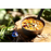 A camping bowl of Happy Yak Chicken Orzo Freeze Dried Soup is shown heated up by a camp stove torch on a tree stump outdoors.