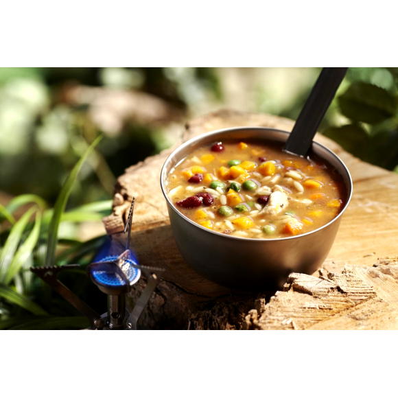 A camping bowl of Happy Yak Chicken Orzo Freeze Dried Soup is shown heated up by a camp stove torch on a tree stump outdoors.