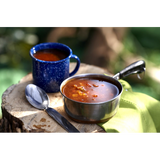 A stainless steel camping pot full of Ranchero Freeze Dried Soup with a white spotted navy cup of the prepared soup, on a tree stump in the woods.