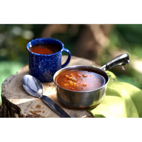 A stainless steel camping pot full of Ranchero Freeze Dried Soup with a white spotted navy cup of the prepared soup, on a tree stump in the woods.