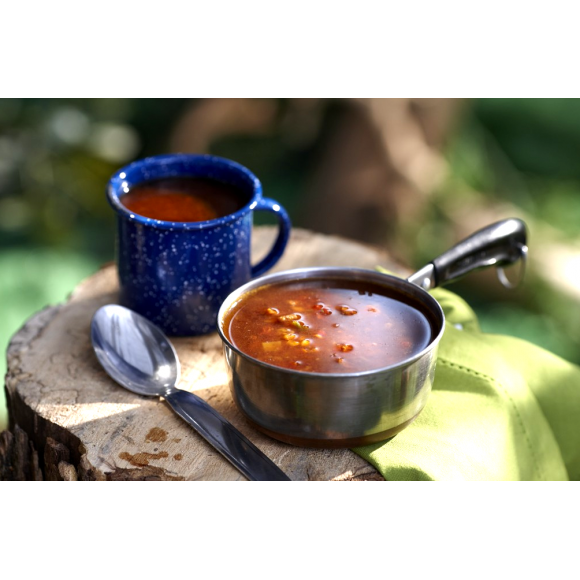 A stainless steel camping pot full of Ranchero Freeze Dried Soup with a white spotted navy cup of the prepared soup, on a tree stump in the woods.