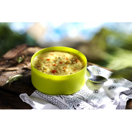 A lime green bowl of the boiled and mixed freeze dried Happy Yak Coconut Thai Soup with a napkin and camping spoon on top of a tree log.