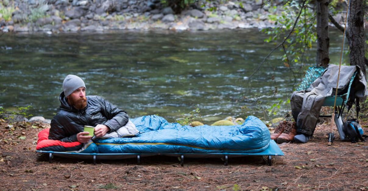 A fisherman drinking a hot cup of tea camping out near a river in the fall wearing laying on a Thermarest Regular Size LuxuryLite Mesh Cot in a blue sleeping bag. The mans fishing gear and waterproof overalls are shown on a chair beside him.